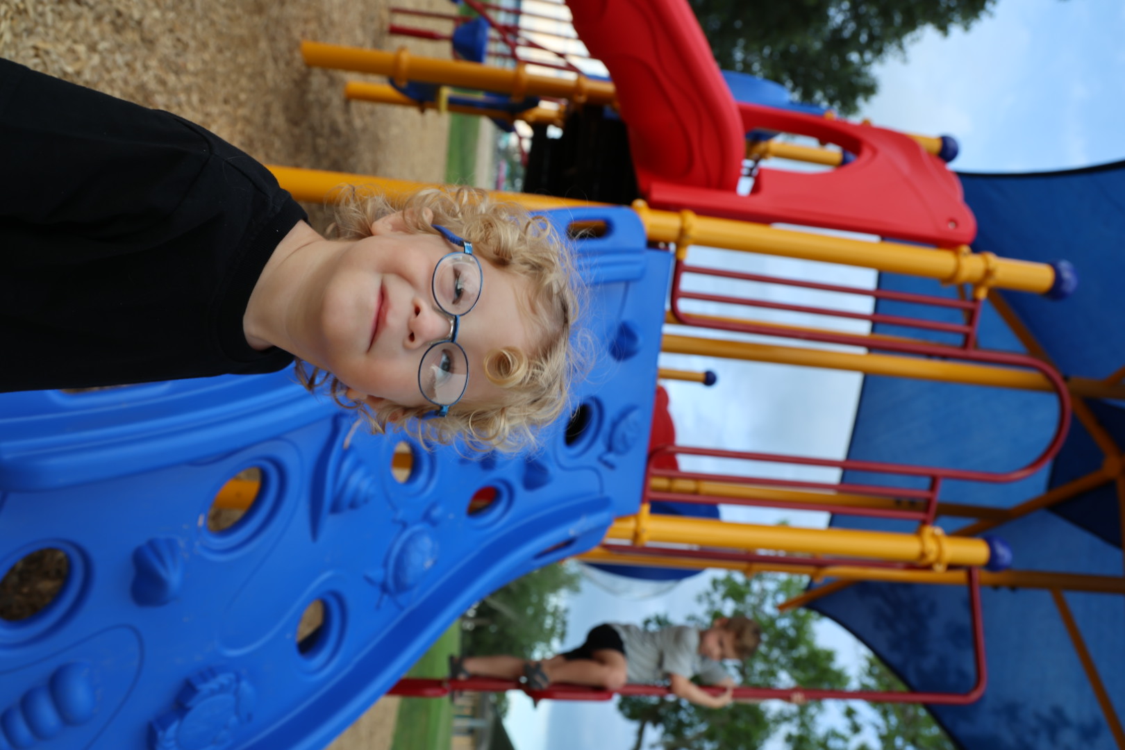 Curly-haired boy in blue glasses smiling in front of a red and blue play structure