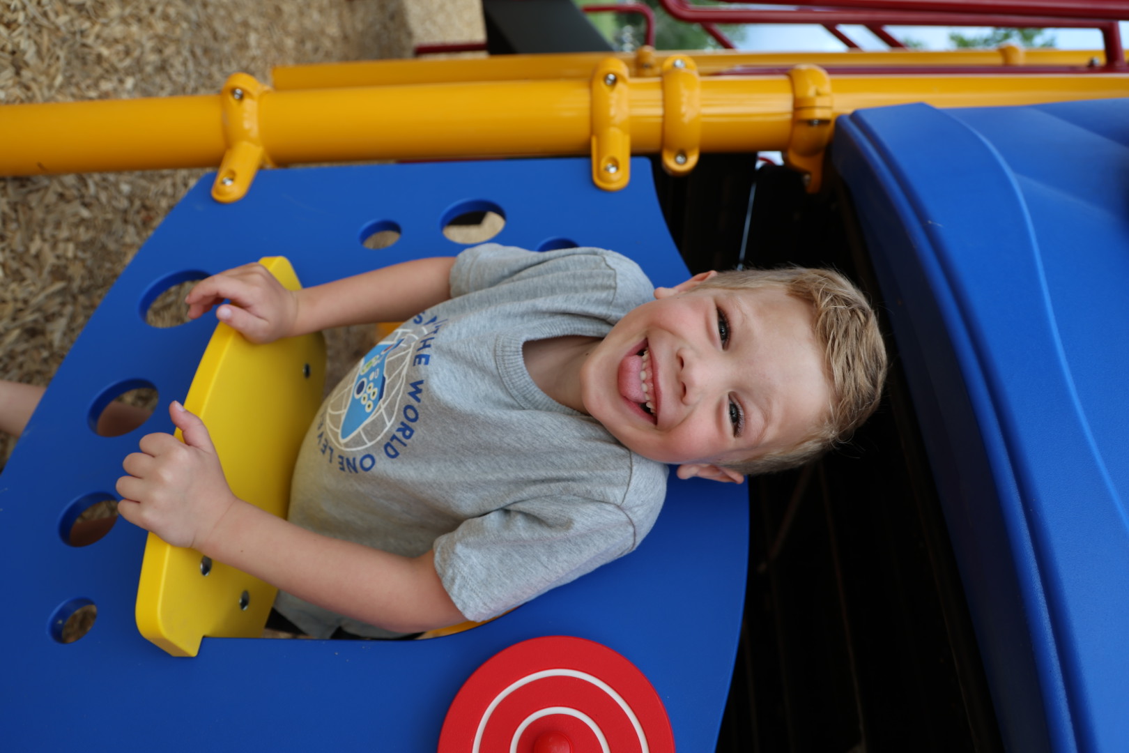 Boy in a gray tee leaning out of a blue playhouse window, tongue out and grinning