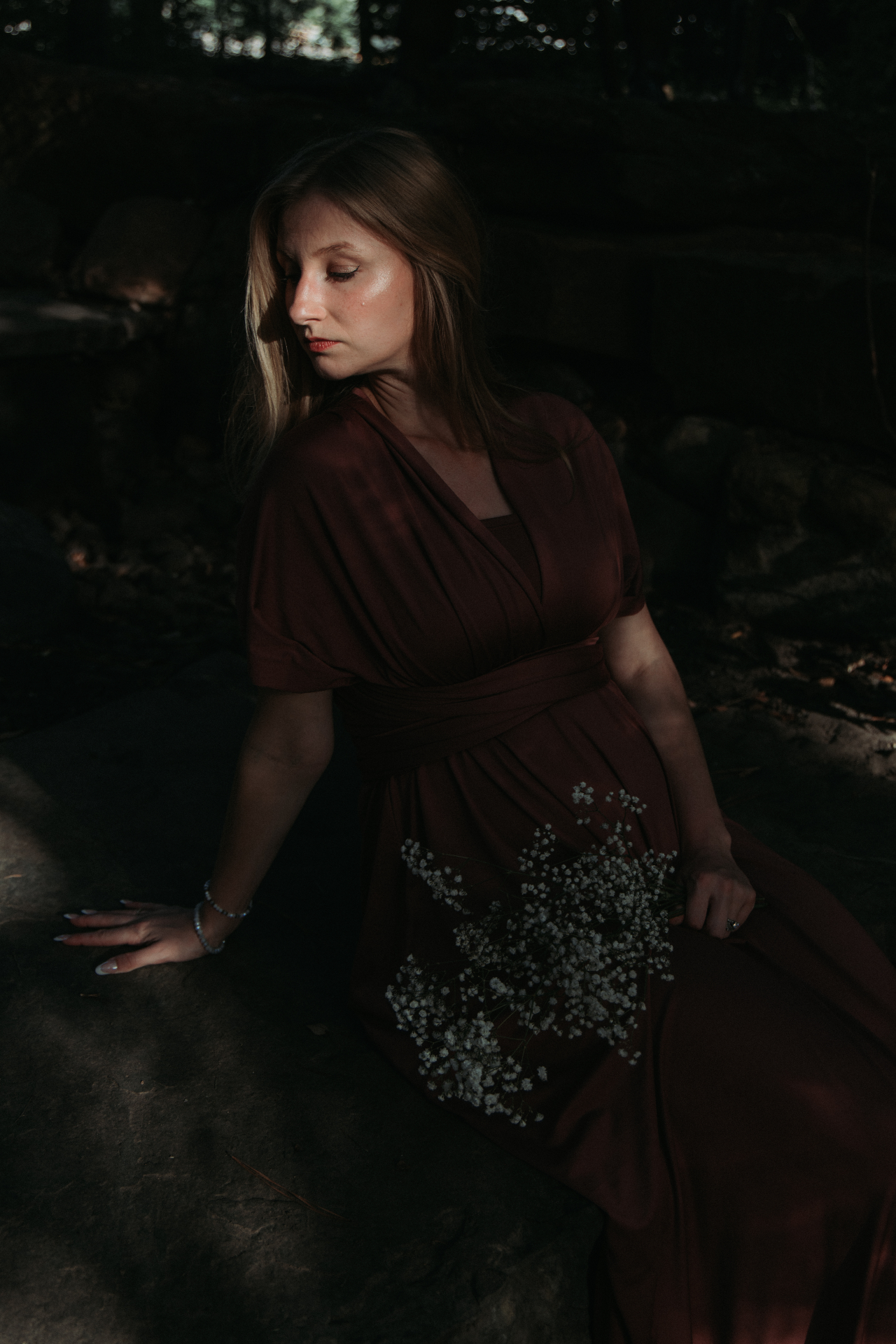 Naomi seated on stone in low light, baby's breath across her lap