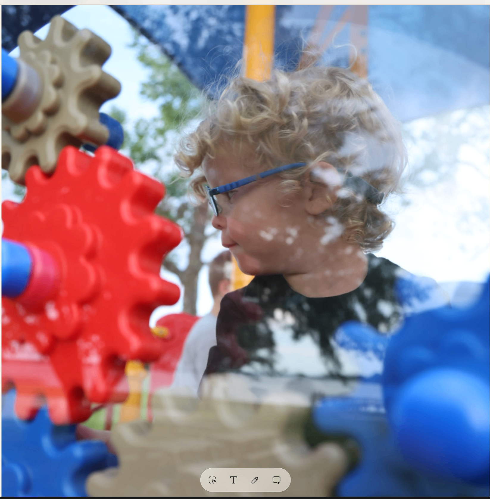 Curly-haired boy in blue glasses turning a red gear on the playground panel