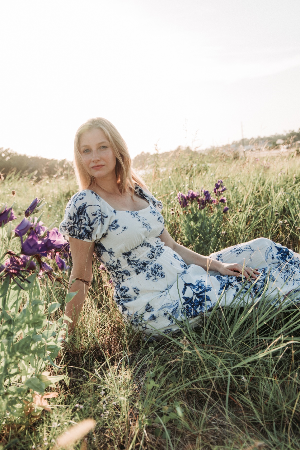 Naomi in a blue floral dress reclining in tall grass at golden hour