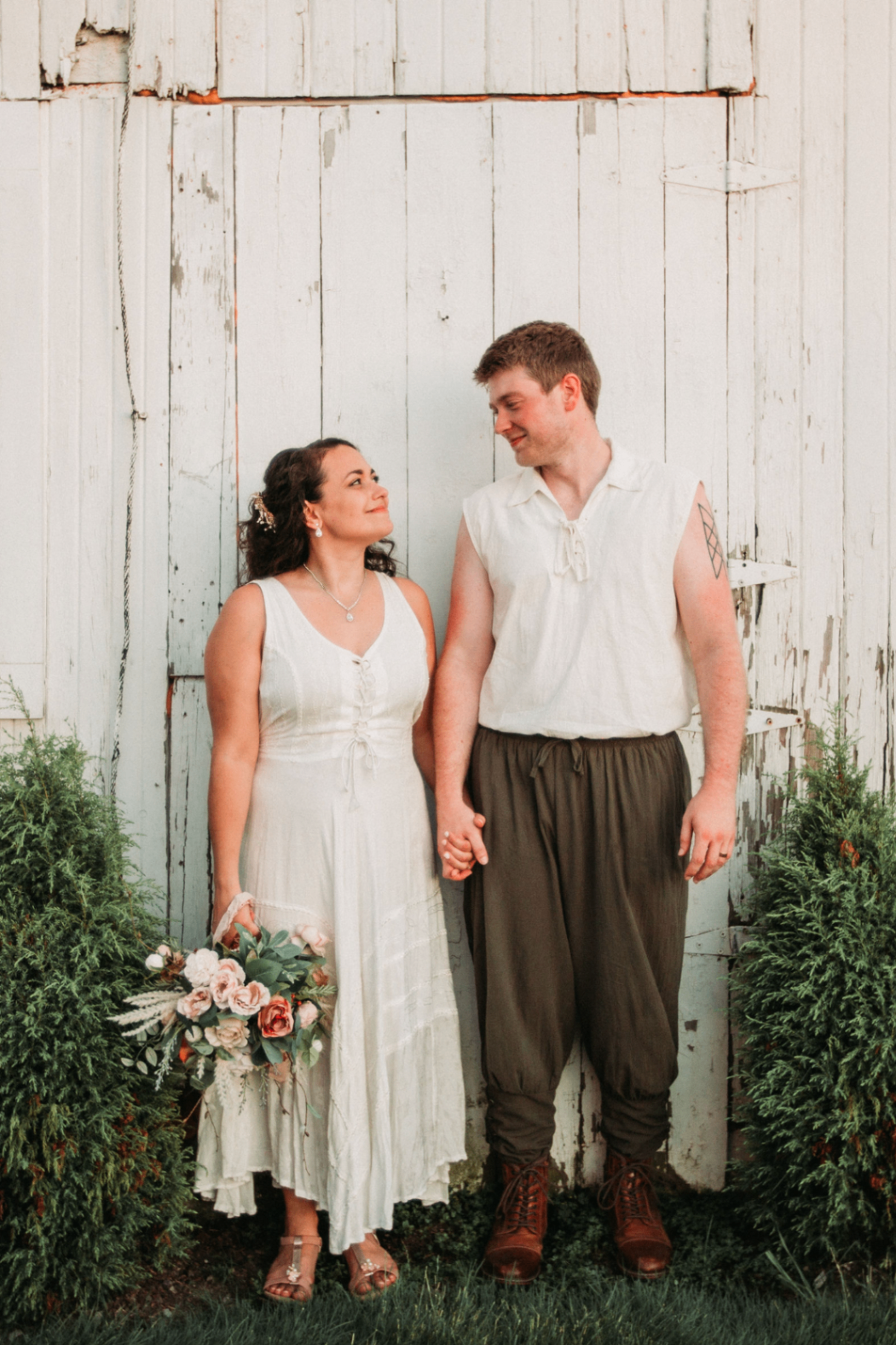 Jessie and Kurtis holding hands between cedar shrubs in front of weathered white barn doors