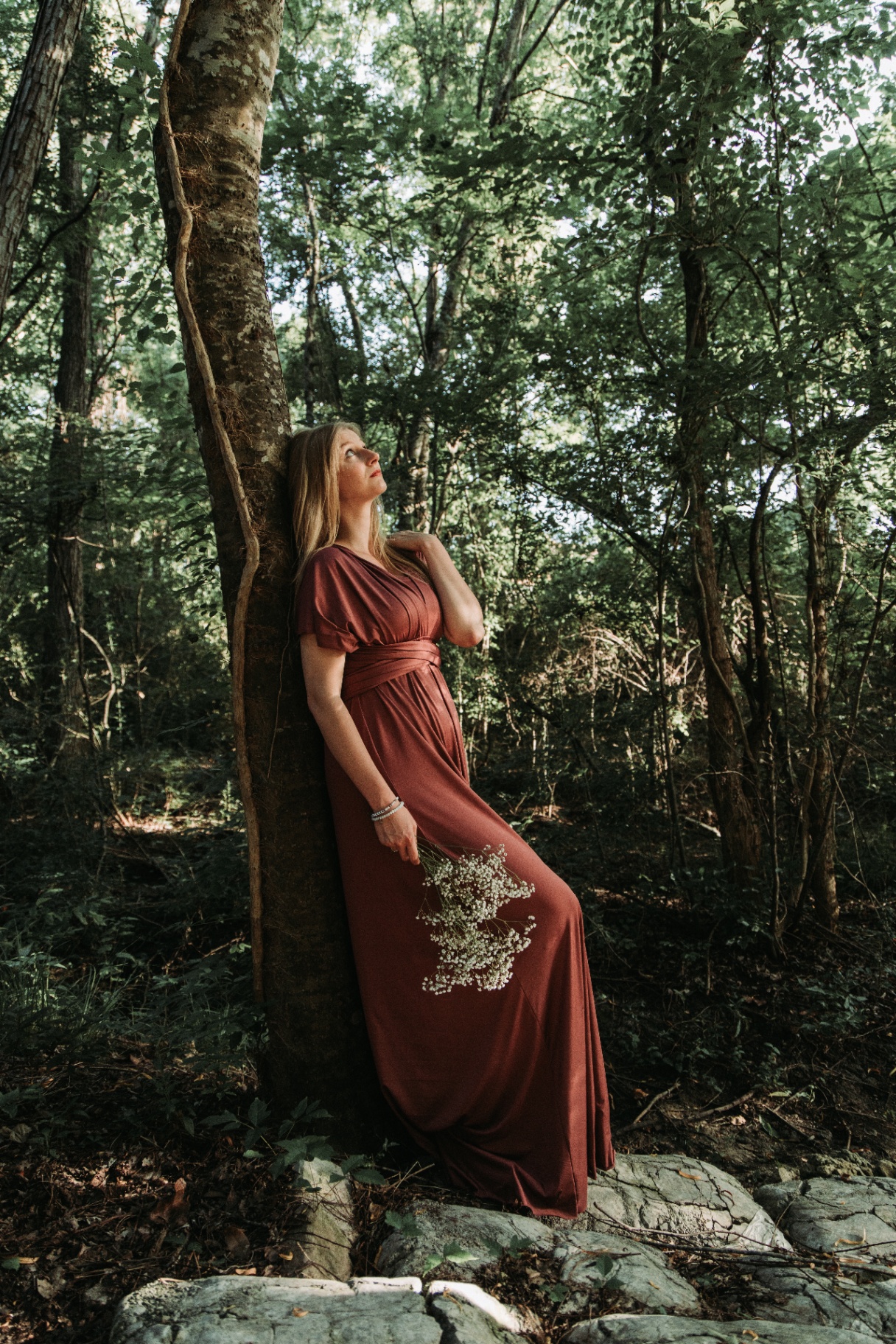Naomi leaning against a slender tree in a rose gown, deep in the woods