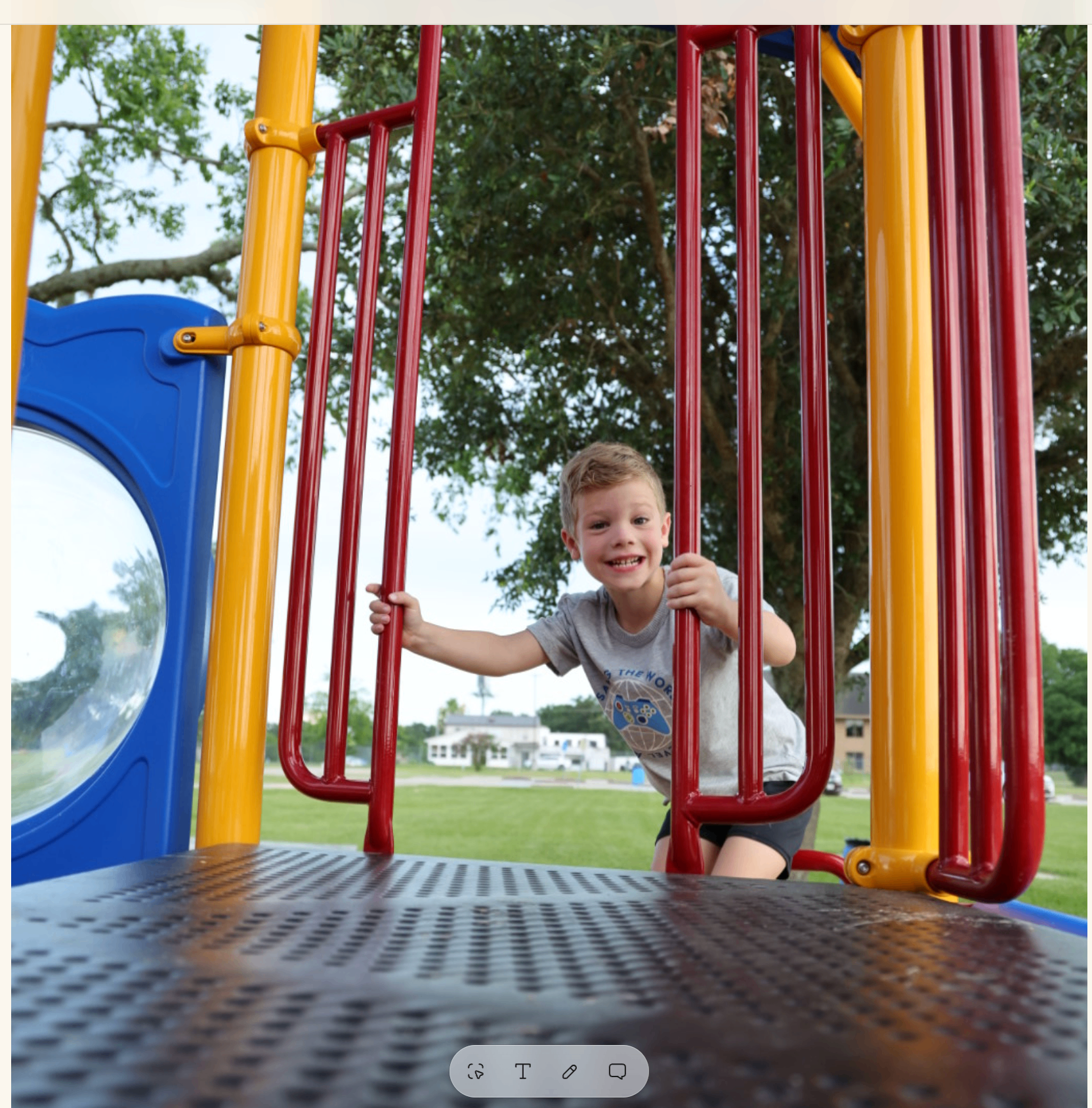 Boy peeking through red playground bars with a wide grin, oak trees behind him