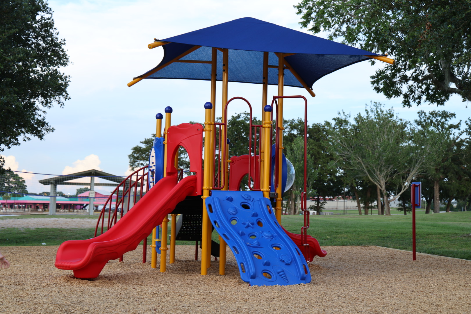Wide view of the red, blue, and yellow playground with a blue shade canopy under summer trees