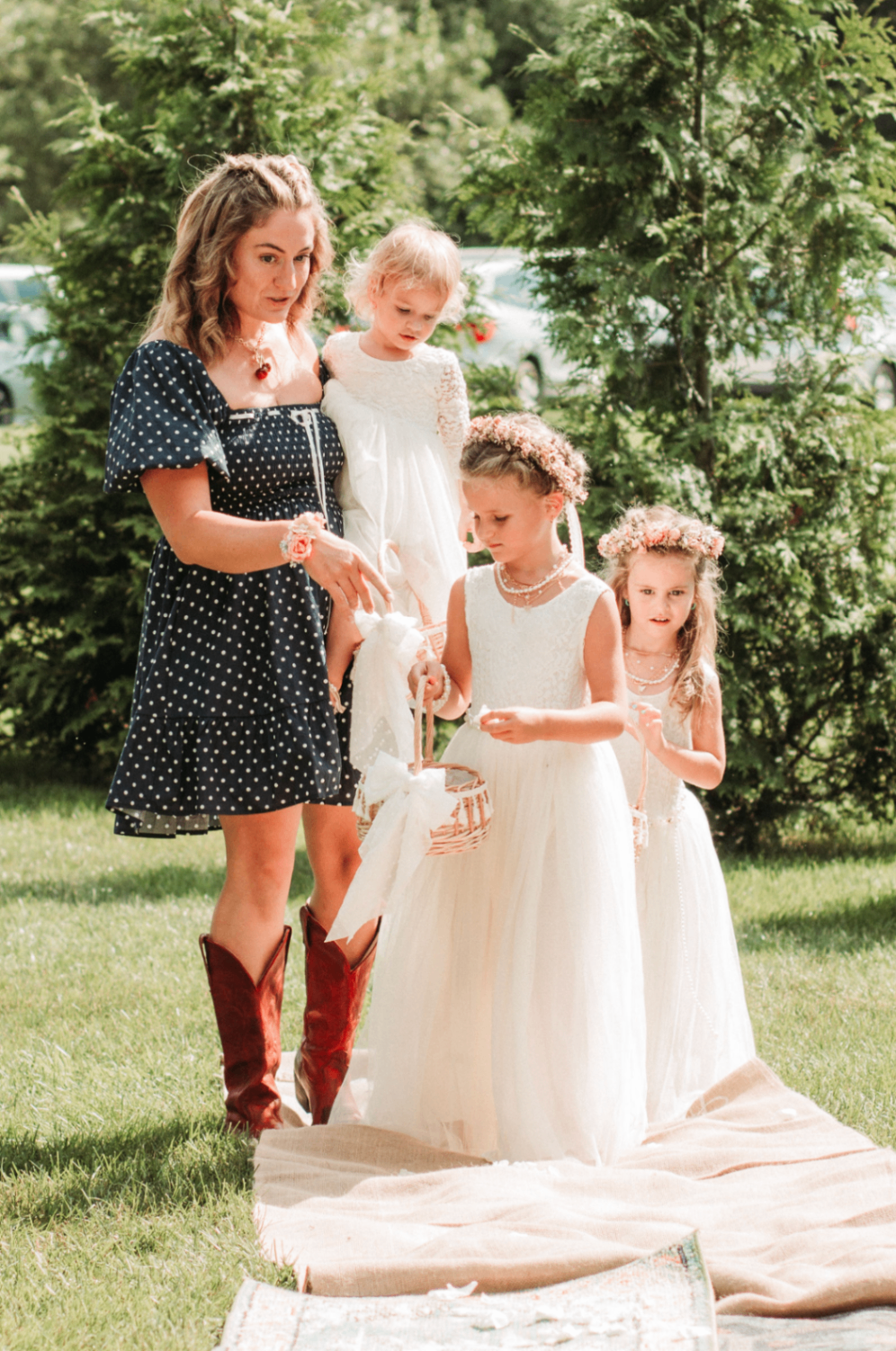 Flower girls holding baskets with a woman in polka dots and red boots on the grass